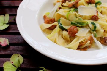 An image of Courgette Pasta with Mushrooms on a white plate standing on a wooden table. There are colorful blooms around the plate.