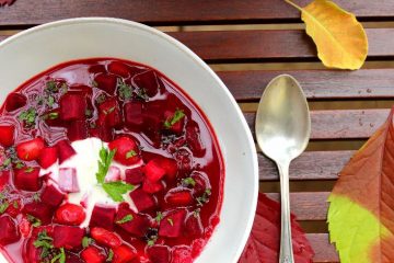 Beetroot Soup Borscht in a white bowl. There is a spoon and a white vase with blooms next to the bowl. Find out about health benefits of beets.