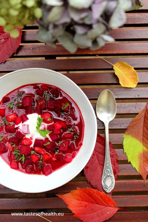 Beetroot Soup Borscht in a white bowl. There is a spoon and a white vase with blooms next to the bowl. Find out about health benefits of beets.