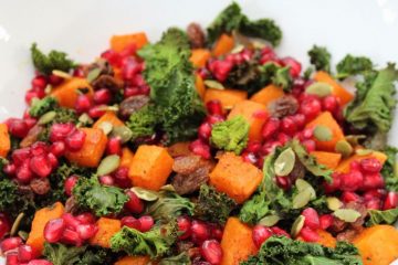 Roasted Kale Salad with Spiced Roasted Pumpkin in a white bowl. There is a pomegranate and a beige cloth behind the bowl.