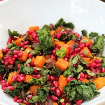 Roasted Kale Salad with Spiced Roasted Pumpkin in a white bowl. There is a pomegranate and a beige cloth behind the bowl.