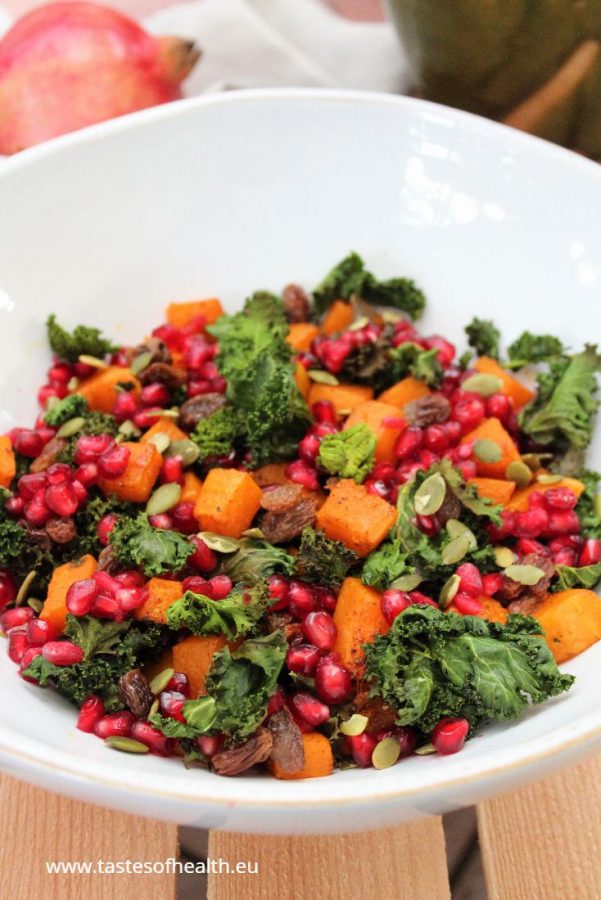 Roasted Kale Salad with Spiced Roasted Pumpkin in a white bowl. There is a pomegranate and a beige cloth behind the bowl.