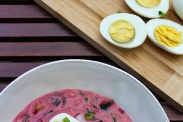 An image of Cold Beet Soup (Chlodnik Recipe With Beet Leaves) in a white bowl. There are cut eggs and chopped chives and radishes on a wooden board next to the bowl.