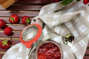 Low Sugar Strawberry Jam Recipe With Pectin - an image of an open jar with strawberry jam seen from above. There are a few strawberries, red roses and a spoon next to the pot. The jar is standing on a white and beige checkered cloth.