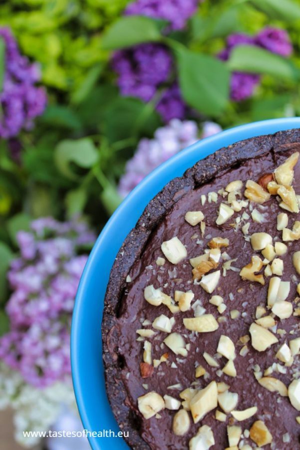 An image of Double Chocolate Tart with Chocolate Fudge and Gluten Free Crust set on a blue plate. In the background there are bushes with blue and purple blooms.