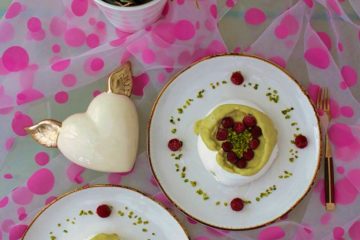 An image for the post: Vegan Meringue Recipe. Two aquafaba meringues with pistachio cream and raspberries arranged in a shape of hearts. The meringues are on white plates. There is also a cream heart with golden wings next to the plates and a piece of fabric with pink dots.