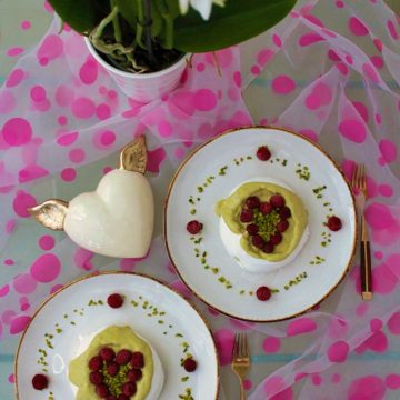 An image for the post: Vegan Meringue Recipe. Two aquafaba meringues with pistachio cream and raspberries arranged in a shape of hearts. The meringues are on white plates. There is also a cream heart with golden wings next to the plates and a piece of fabric with pink dots.