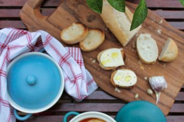 An image of Roasted Tomato and Red Pepper Soup with Mozzarella Bruschetta in a white bowl. There are two blue lids, slices of bruschetta with mozzarella on a wooden board, a plant with red fruit and a white and red cloth next to the soup.