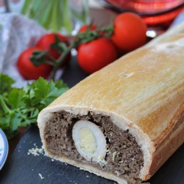 An image of Vegetarian Pork Pie with Hot Water Crust Pastry. The pie is cut open so you can see its inside: mushrooms and eggs. The pie is on a slate plate and there are red tomatoes in the background.